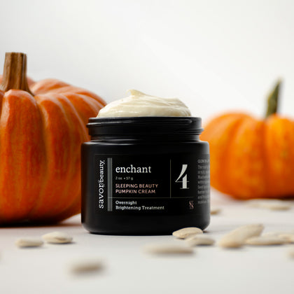 A black jar of Savor Beauty Enchant Sleeping Beauty Pumpkin Cream with white cream visible on top, styled on a white surface with pumpkin seeds in the foreground and orange pumpkins blurred in the background.