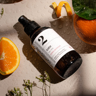 Wet dark spray bottle with white label reading '2' laying on a neutral background surrounded by fresh oranges and greenery.