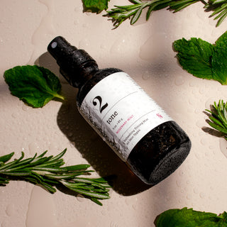 Wet dark spray bottle with white label reading '2' laying on a neutral background surrounded by mint leaves and rosemary sprigs.