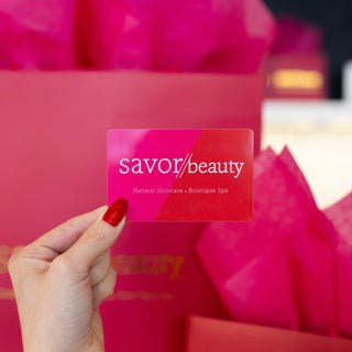Hand holding a Savor Beauty card with pink gift bags in the background