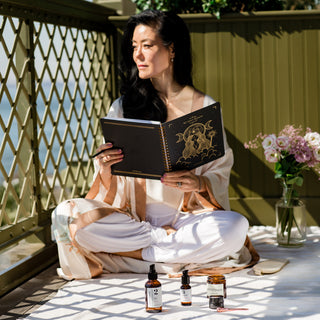 Founder Angela Jia Kim working on her Savor Beauty Planner on a olive green balcony with miscellaneous skincare and spring florals