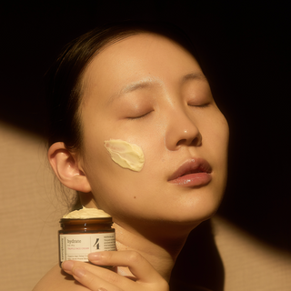 Woman applying cream to her face with a jar of cream in front of her.