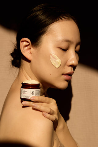 Woman applying cream to her face with a jar of cream in hand against a neutral shadowed background