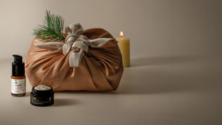 A styled product shot featuring a brown fabric gift bag tied with a beige ribbon and decorated with pine needles, alongside a small amber bottleof pomegranate eye serum and a black jar of caviar collagen eye cream, set against a warm beige background with a lit candle.