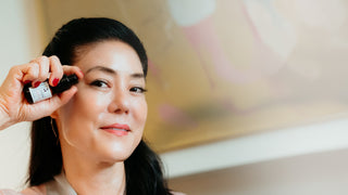 Founder Angela Jia Kim applies pomegranate peptide eye serum from a small amber/black pump bottle. The background is a softly blurred warm-toned interior.